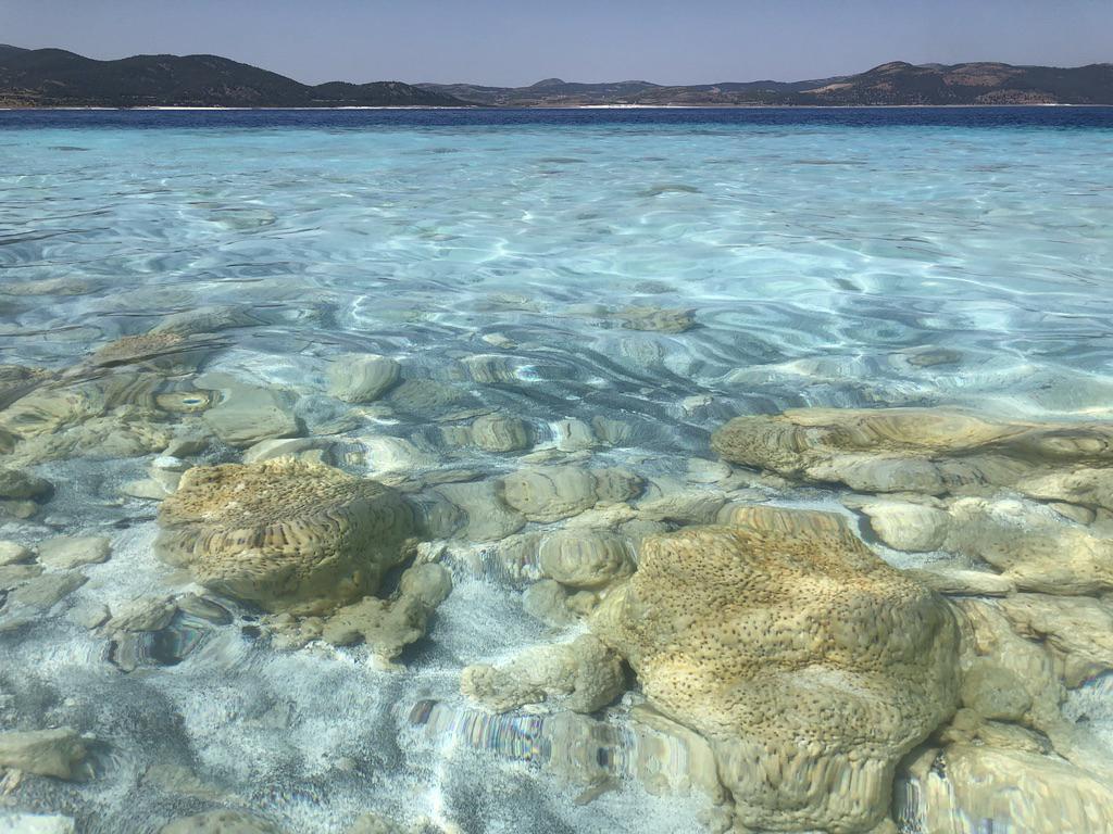 The clear, light blue water of Lake Salda fills most of the frame. Across the lake, hills can be seen on the horizon. Beneath the water are yellowish structures that resemble balls of coral that are almost brain-like.