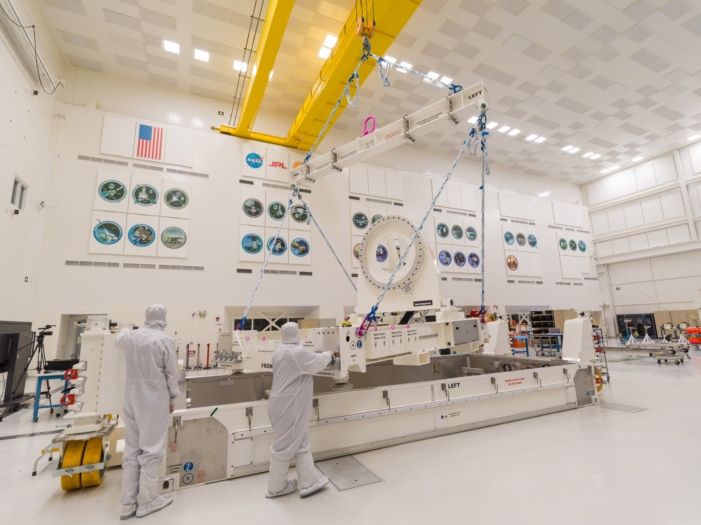 Workers in white protective clothing use a crane to lift and move cream-colored equipment that will be used to help build NEO Surveyor.