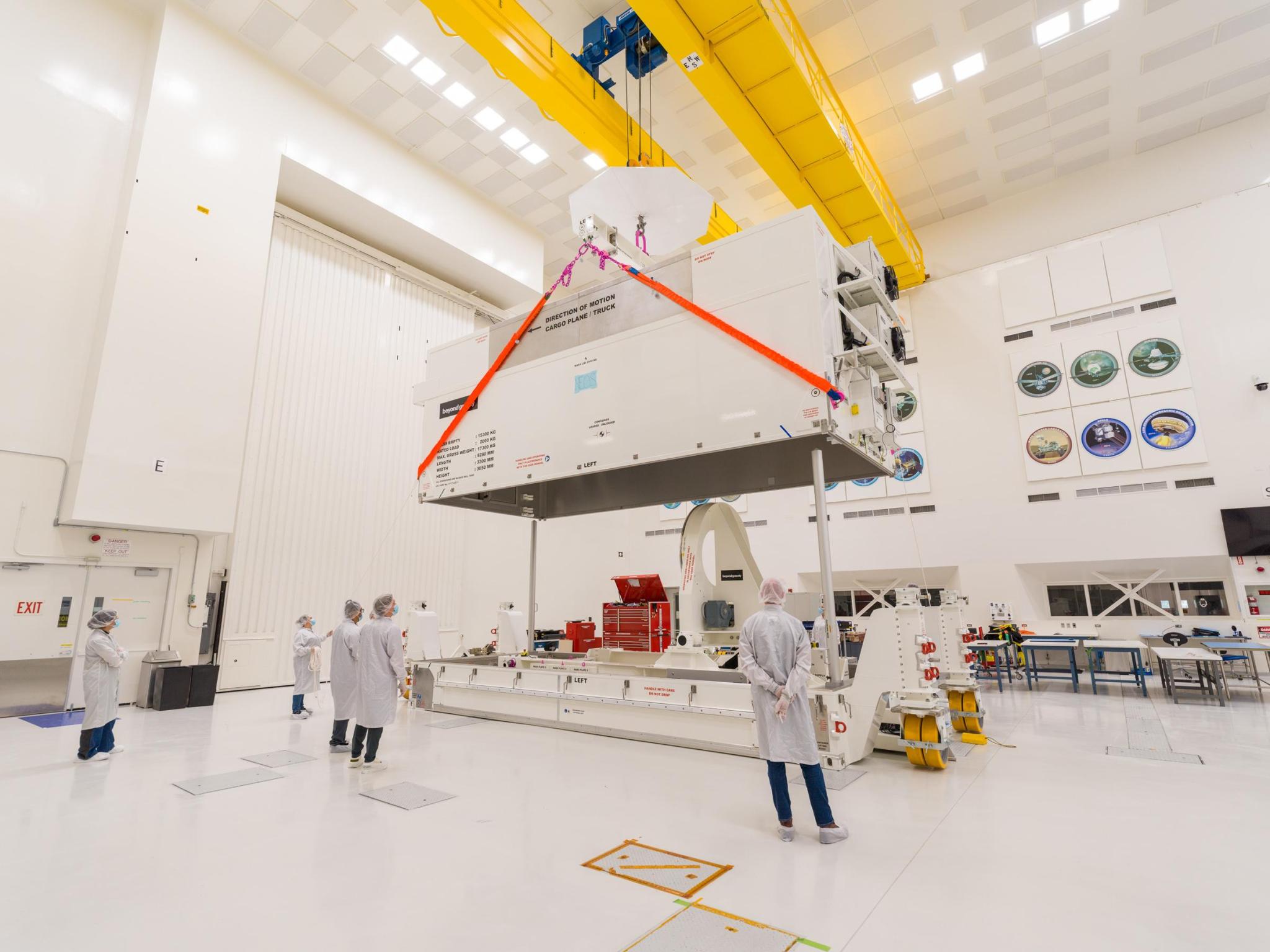 Workers in white protective coats and head coverings stand nearby and watch a yellow crane lift a cream-colored lid protecting equipment to be used during construction of NEO Surveyor.