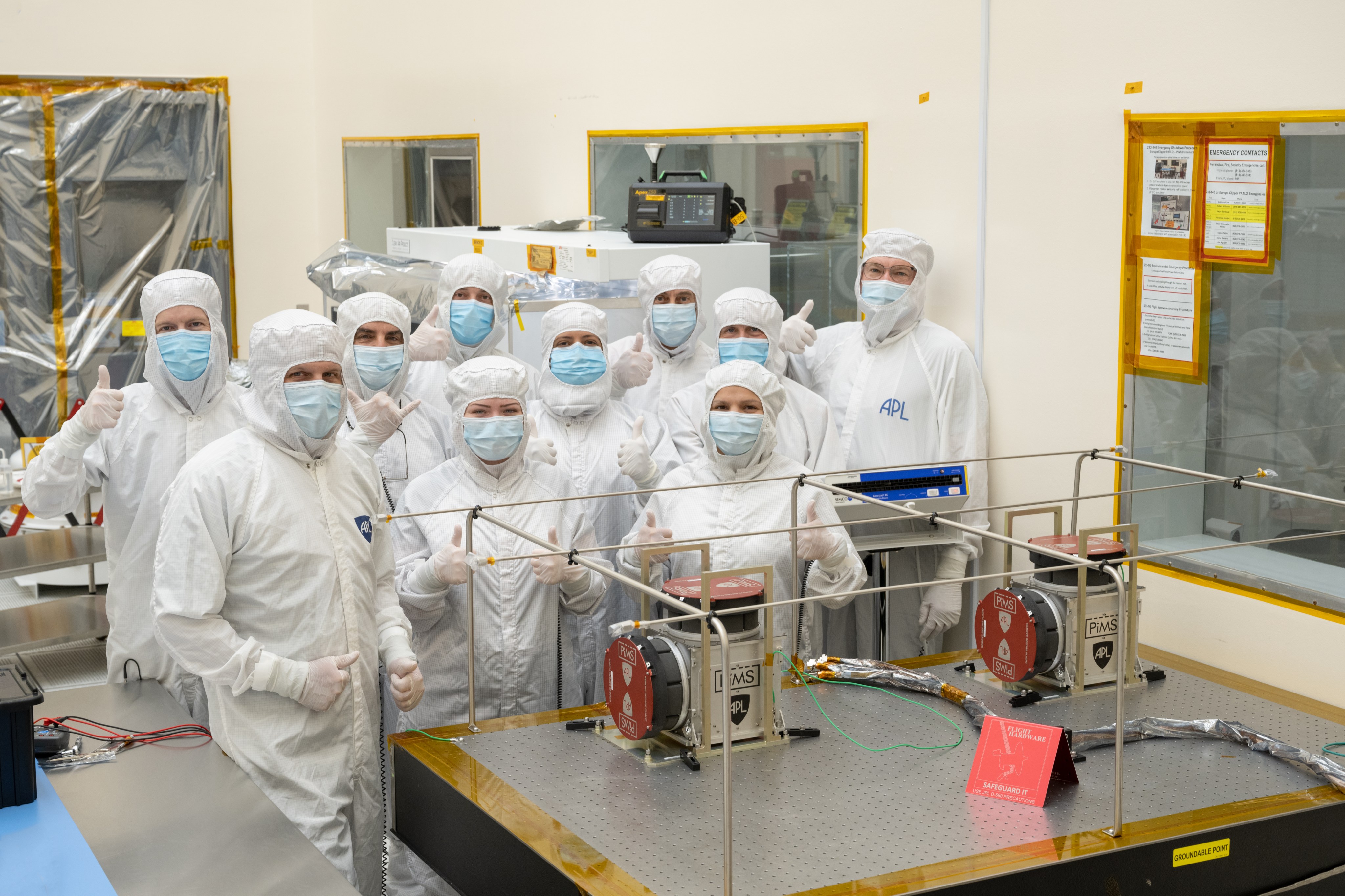 Ten workers in white protective clothing and blue face masks stand behind a table with a spacecraft instrument on it and give a thumbs up.