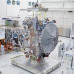 A large silver colored dish-shaped antenna hangs on the side of the main Europa Clipper spacecraft body on a work stand. Workers are the floor nearby.