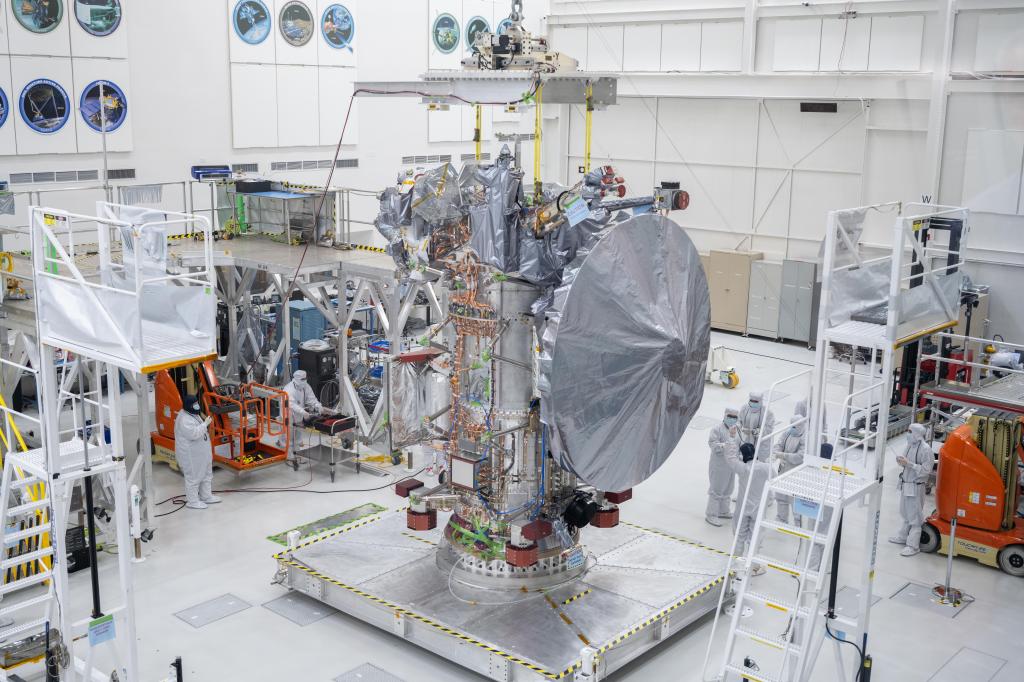 A large silver colored dish-shaped antenna hangs on the side of the main Europa Clipper spacecraft body on a work stand. Workers are the floor nearby.