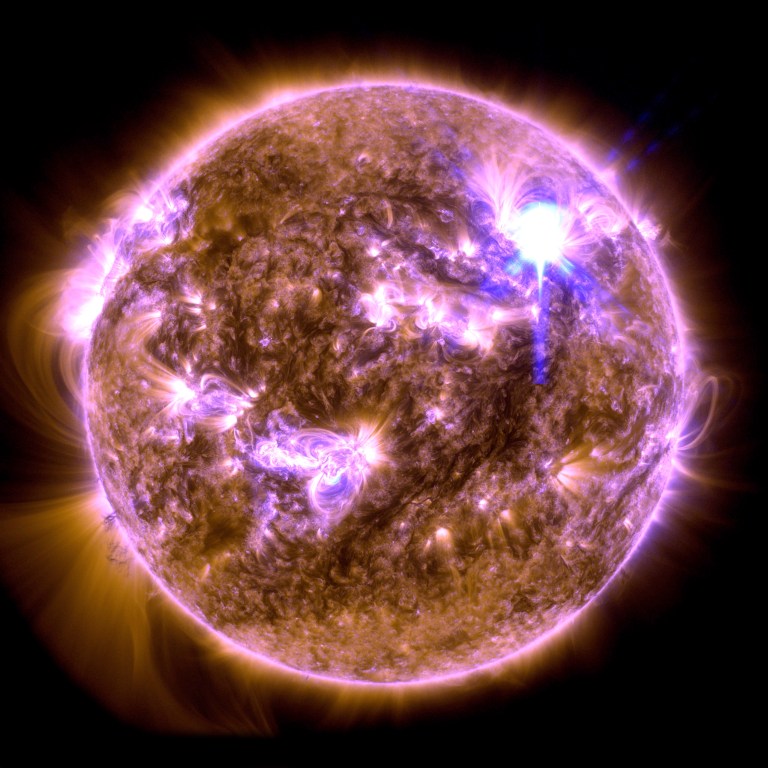 The Sun appears as a mottled orange orb, with several bright regions of swirling plasma appearing in pink and purple. A bright white flash of light appears in the upper right.