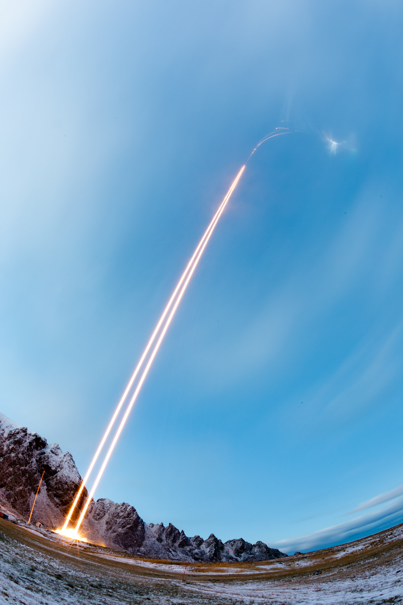 A time-lapse photo shows a rocket launching into the sky, leaving a bright, arcing trail against a backdrop of mountains and a clear blue sky. Snow-covered ground and scattered clouds are visible, with the horizon stretching below.