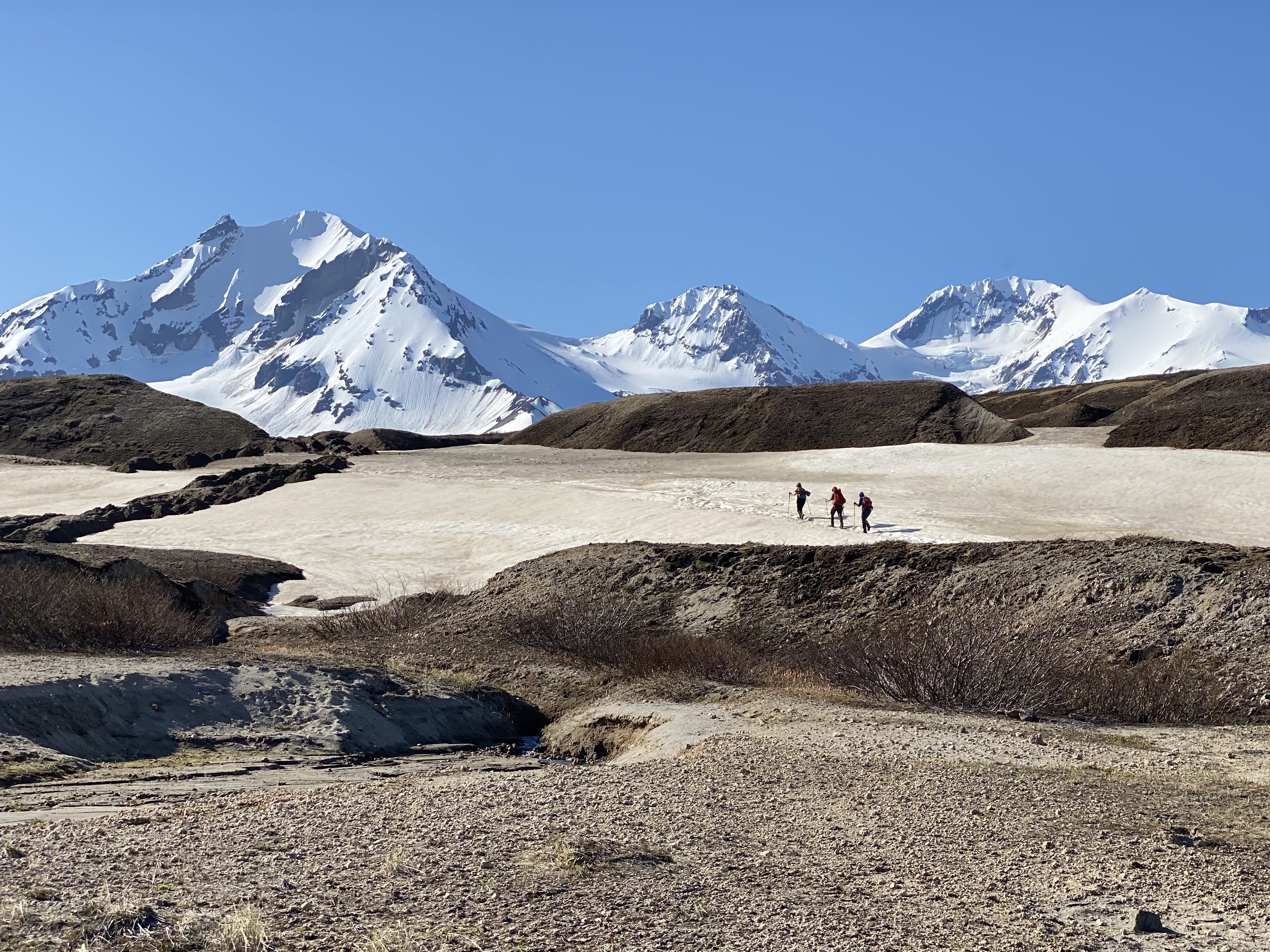 Three people, wearing large backpacks, trek across a snow field between hills of dark rubble. In the background: steep, snow-covered mountains under a blue sky.