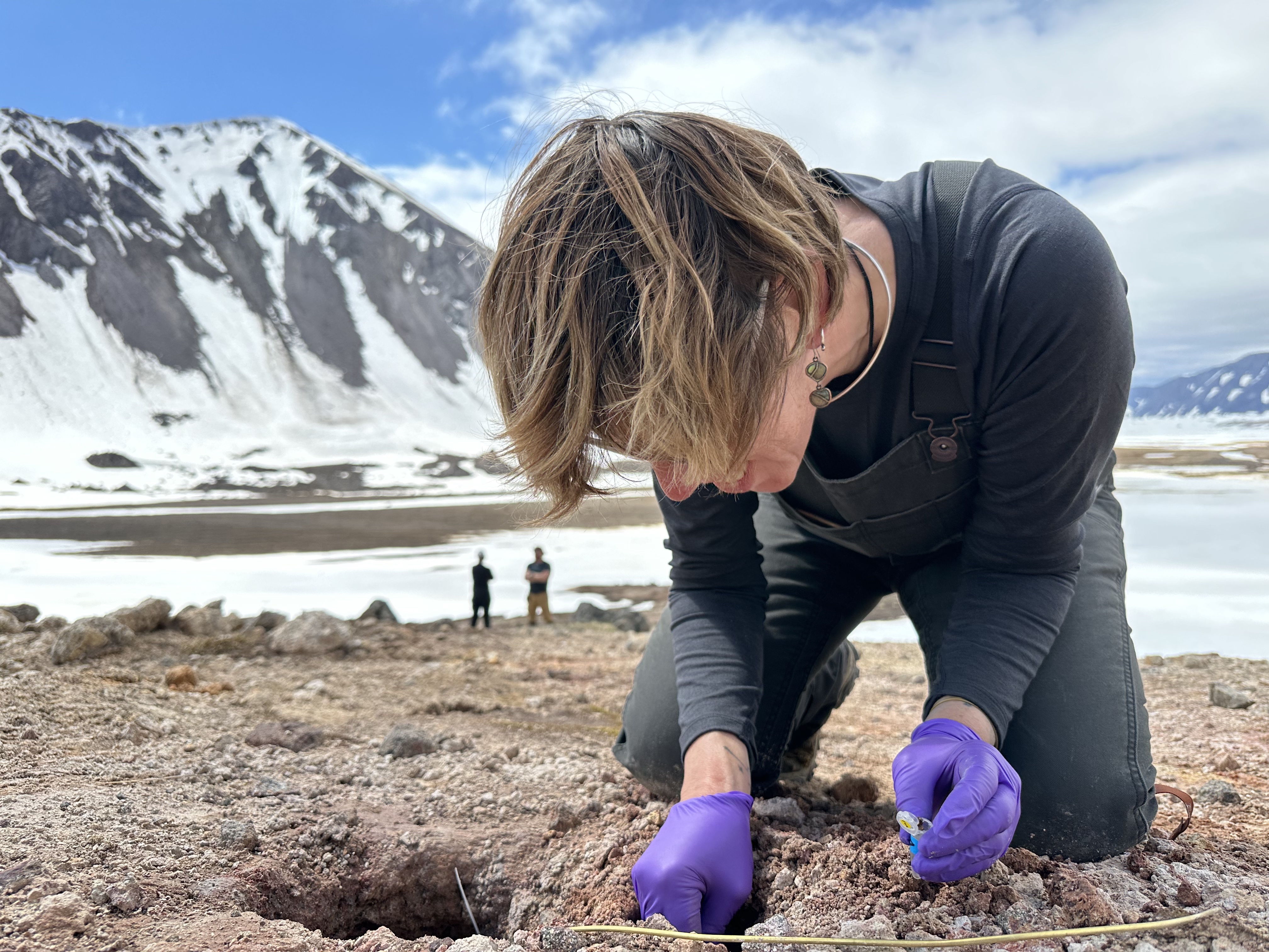 Person kneeling on reddish-brown, rocky ground, near a small hole, with a steep, snow-patched mountain in the background. They are wearing purple nitrile gloves and holding a tiny, open vial in one hand while digging with the other. A golden wire stretches across the dirt and into the hole in the ground.