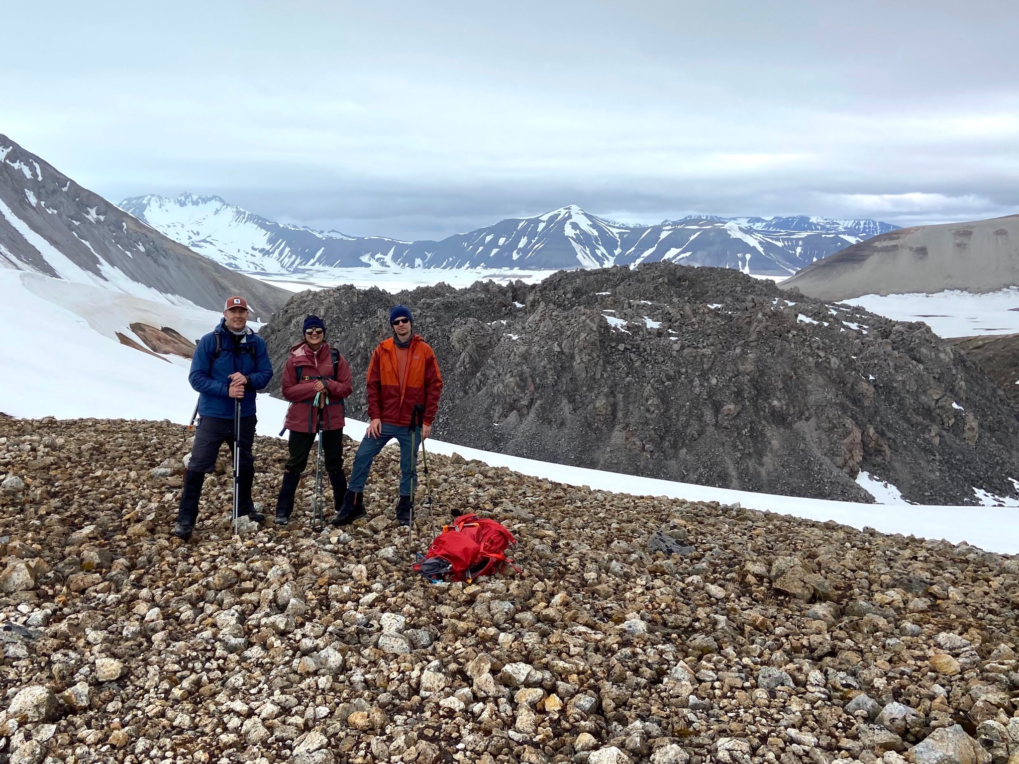 Three people, dressed for outdoor work, on a rocky hill in front of a mountainous landscape under an overcast sky. In the middle distance is a huge, dark-colored pile of rubble, shaped like a low dome.