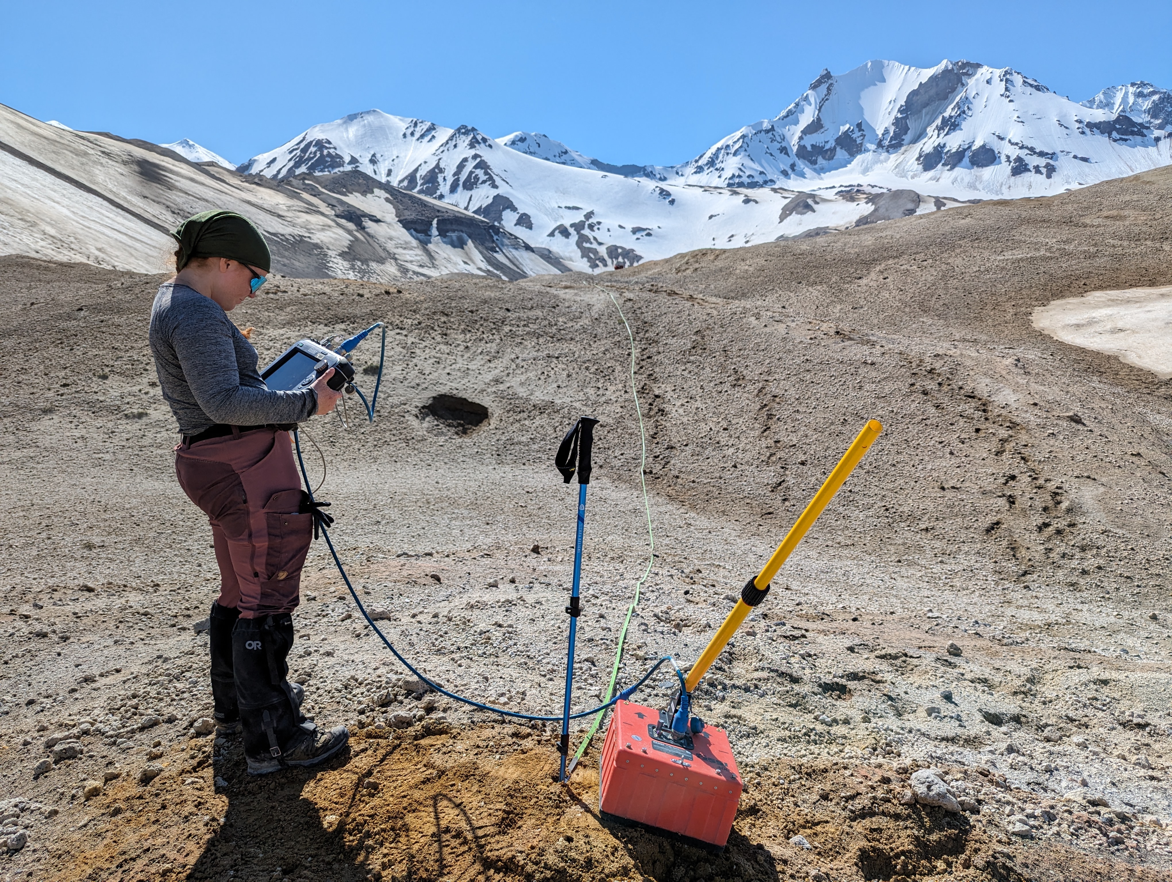 Person holding a bulky computer readout attached via a thick cord to a red plastic box with a push handle, on an expanse of beige volcanic ash, with snowy mountain peaks in the background. A tape measure, anchored to the ground with a trekking pole near the red box, extends over a hill into the distance.