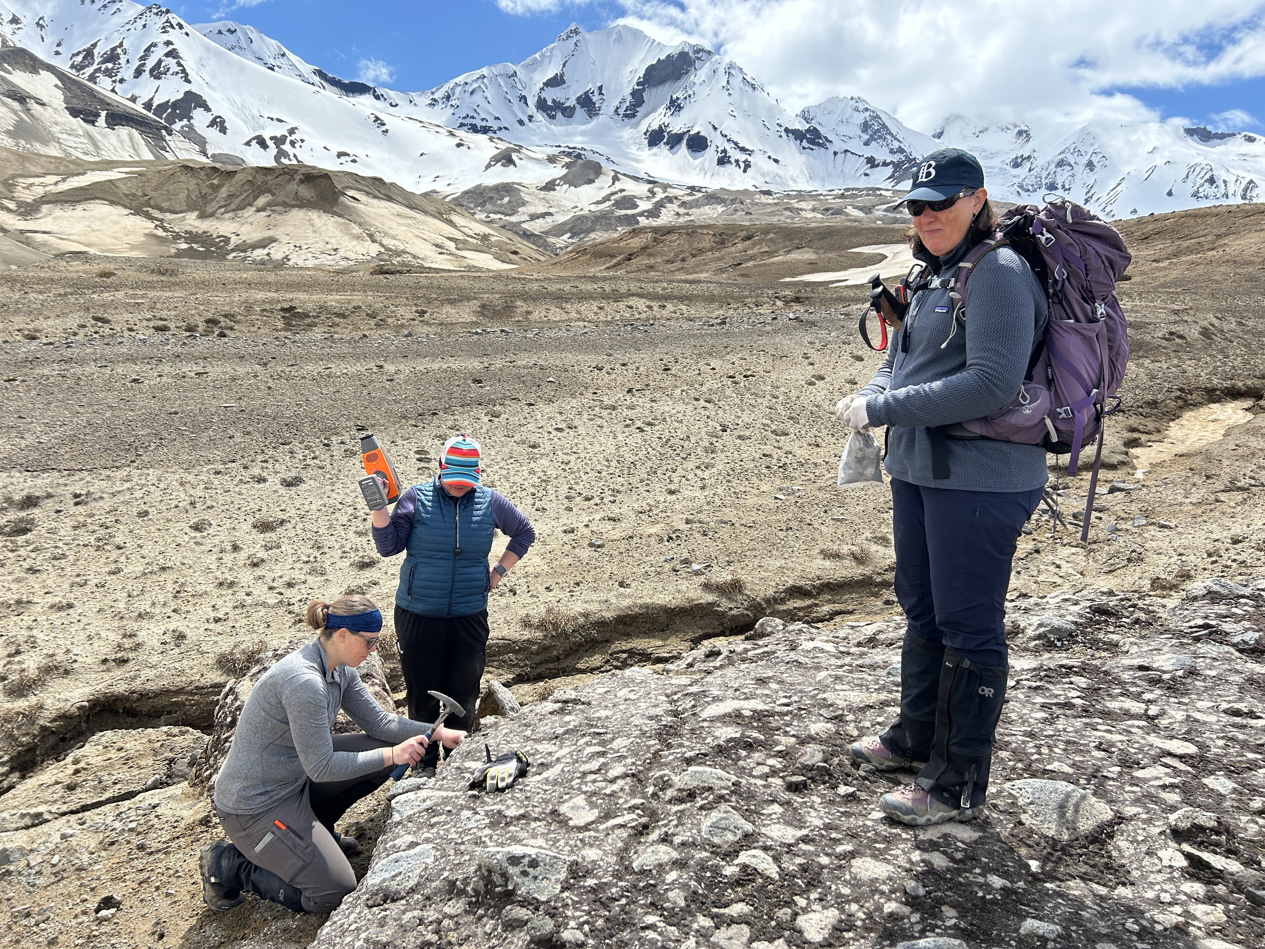 Three people on a barren, rocky landscape with hills of grey ash and snow-covered mountains in the background. The researcher on the left kneels and raises a rock hammer, about to collect a sample. Nearby, another scientist props a portable spectrometer up on her shoulder in between uses-- the spectrometer resembles a large, orange and grey blow dryer. The third scientist holds a bag of rock samples and looks at the camera. She has a large pack on her back and hiking poles under her arm.