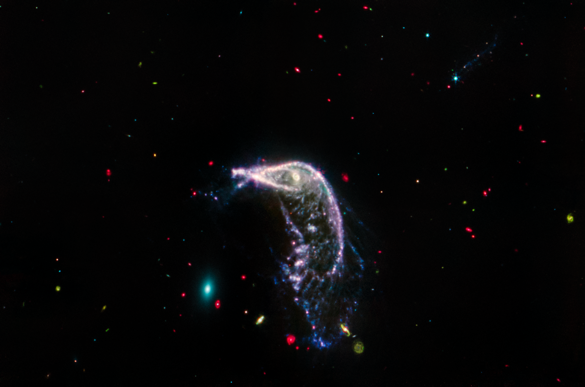 Two interacting galaxies known as Arp 142 in a horizontal image taken in mid-infrared light. At left is NGC 2937, an elliptical galaxy that looks like a tiny teal oval and is nicknamed the Egg. At right is NGC 2936, a distorted spiral galaxy nicknamed the Penguin, which is significantly larger. A beak-like region points toward the Egg, but lies far above it. Where the eye would be is an opaque, almost washed-out pink spiral. This galaxy’s distorted pink, purple, and blue arms create the bird’s beak, back, and tail. The tail, which is closer to the Egg, is wide and layered, like a beta fish’s tail. The Penguin and the Egg appear very separate. The galaxy at top right, PGC 1237172, is barely visible. A brighter slightly larger blue foreground star that is overtop this galaxy has tiny diffraction spikes. Throughout the image are tiny galaxies in bright reds, greens, and blues. The background of space is black.