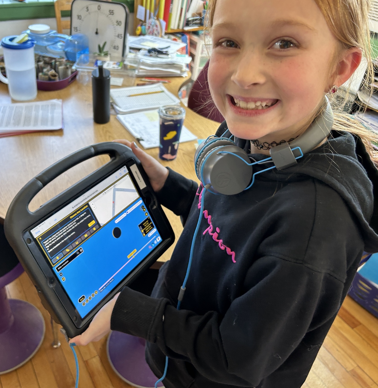 A smiling 8-yo student holds a digital tablet that is displaying the Total Eclipse 2024 Data Story. On the tablet screen, a map shows a red dot at the location selected by the student. Underneath the map, a view of the Sun and Moon shows the Moon almost completely overlapping the Sun, with a sliver of the Sun still visible.