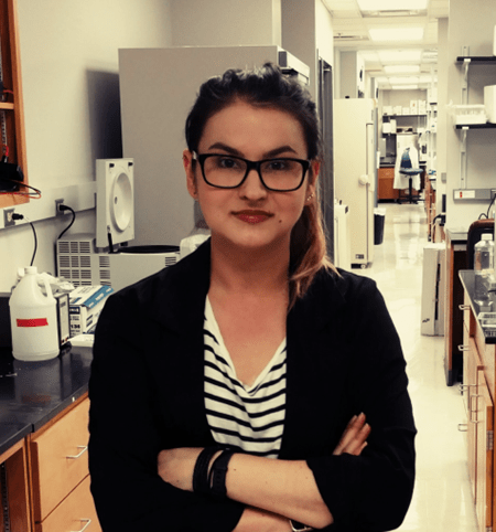 Katarzyna Piekarz, Ph.D. Portrait photo of a young woman wearing glasses and a black lab coat; a laboratory interior is visible in the background.