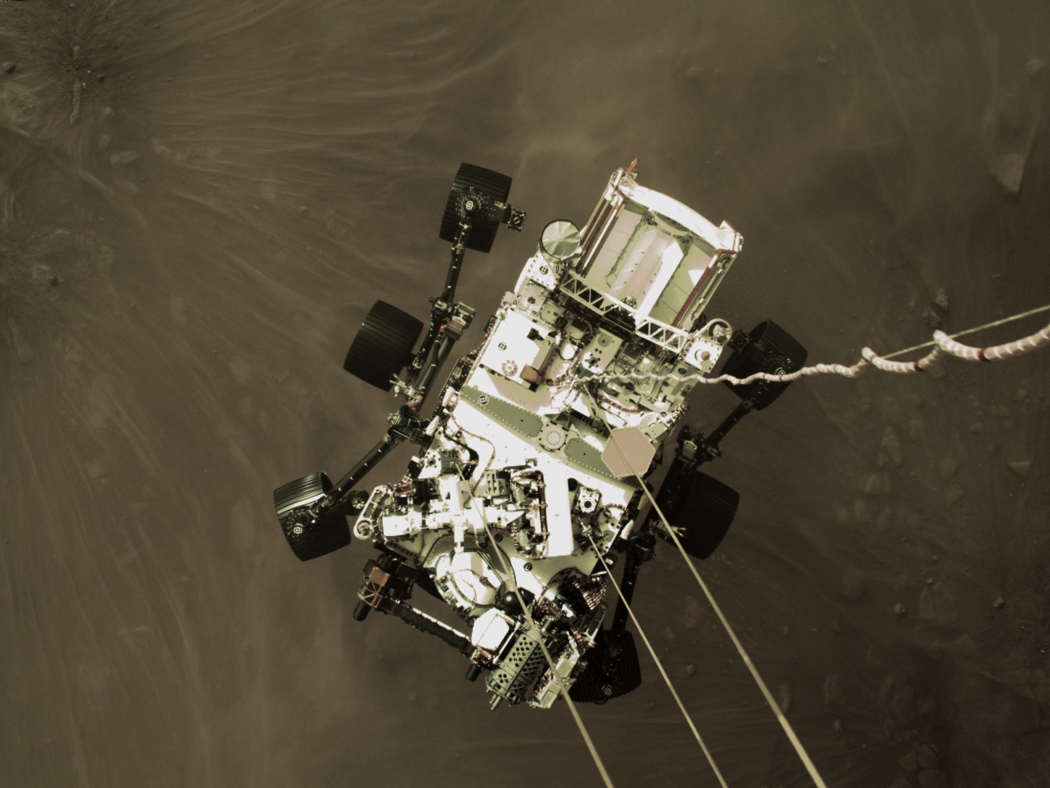 An overhead photograph of a six-wheeled Mars rover, mostly white with black wheels, touching down on the dark brown-gray surface of the planet while still suspended on tethers reaching up to an unseen descent craft hovering above it.