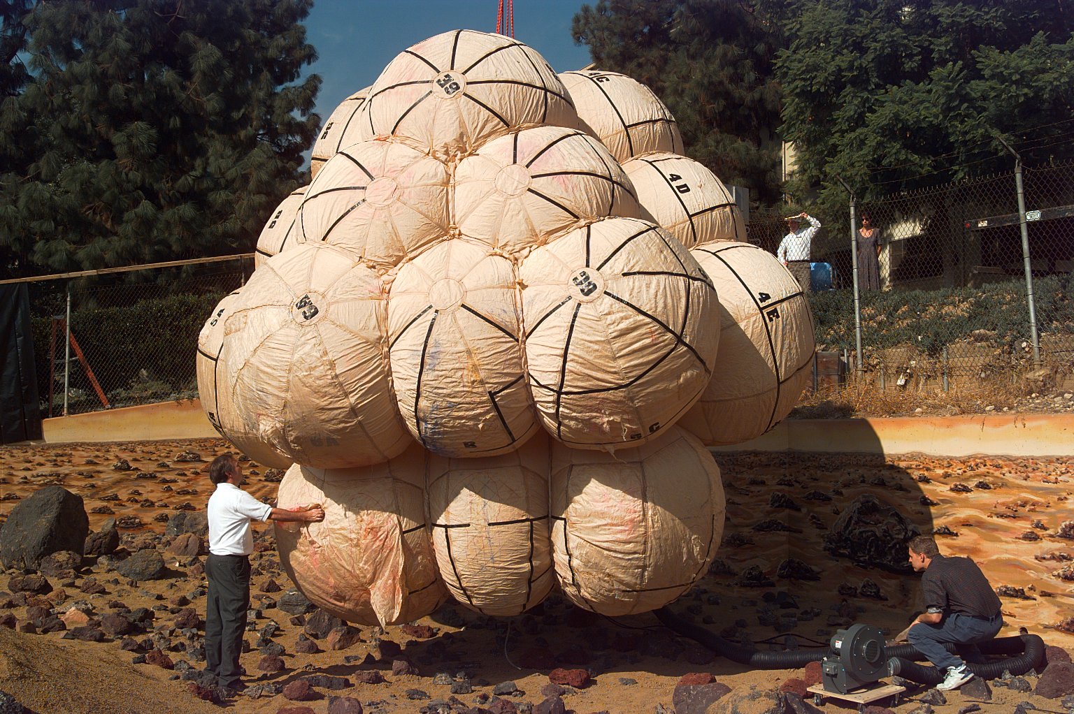 A man dressed in dark slacks and a white shirt stands outdoors at image left, examining a giant airbag composed of large, fabric balloons, each about as large as the man, all attached to each other, with more than a dozen visible. Another man in blue jeans and black shirt, crouches at left, operating what appears to be an air blower, which has a wide black hose extending to the underside of the airbag package.