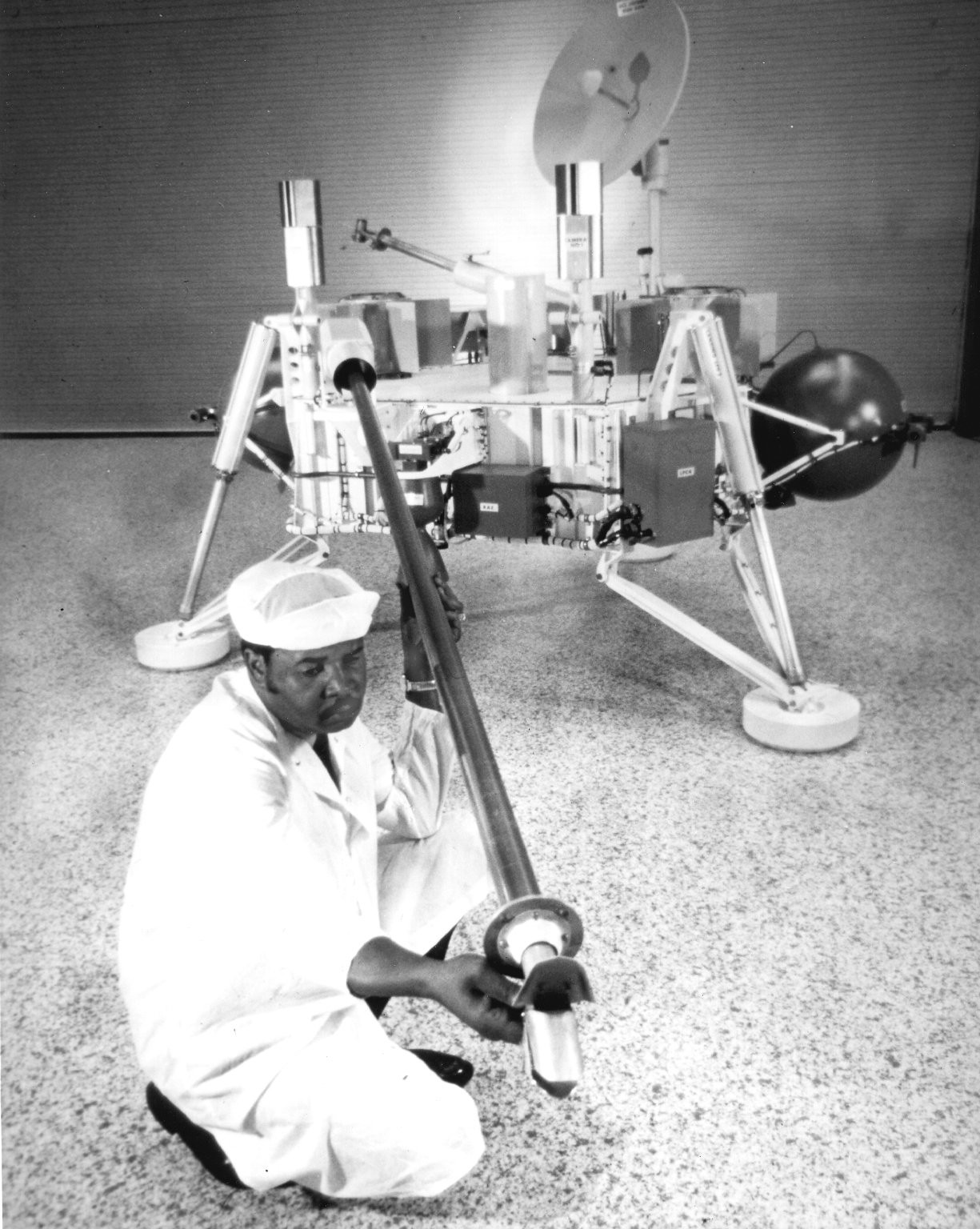 A man dressed in a white work clothes and white head covering examines the end of a long boom arm extending from a squat, three-legged spacecraft with a radio antenna dish perched on its top, inside an otherwise empty work room.