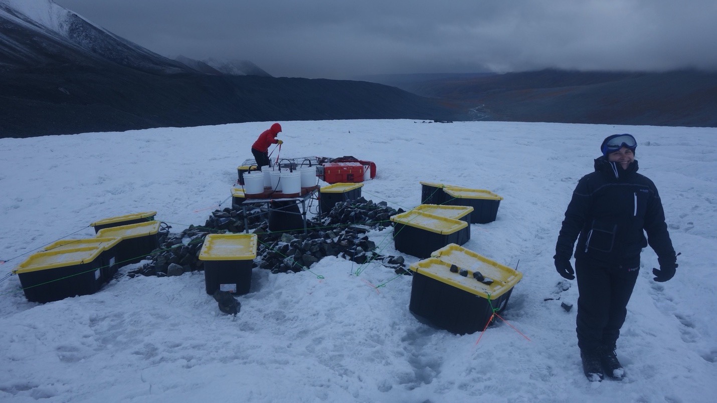 A smiling, bundled-up researcher stands beside an array of heavy-duty containers and equipment spread across a dim, icy landscape. Behind her, another researcher works. In the background, dark mountains rise into misty, heavy clouds.