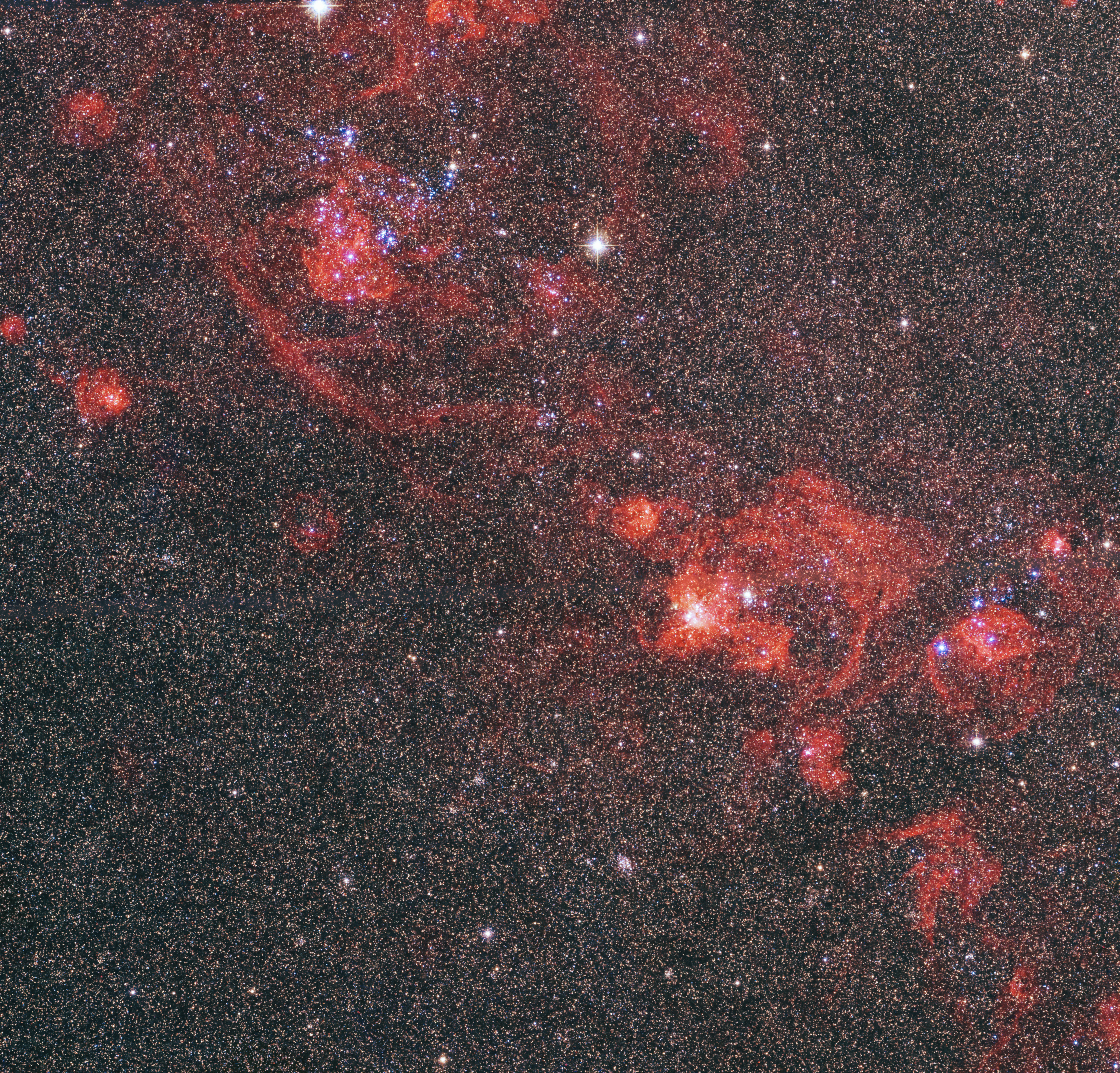 Thousands of distant stars crowd the view against black space. Tendrils of red nebulosity bloom across the image.