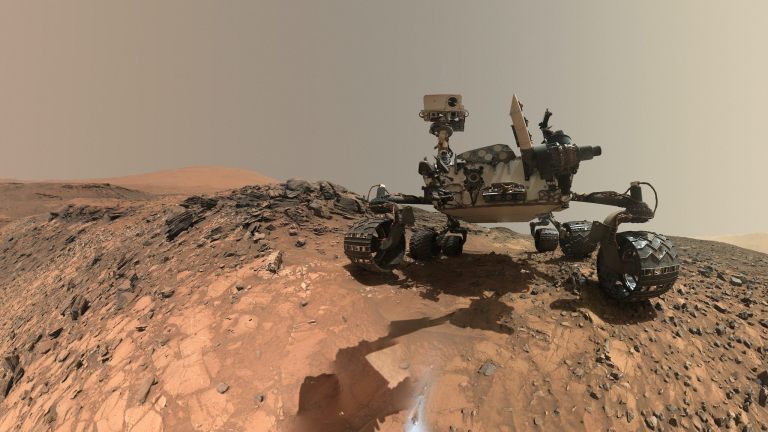 A rover sits on the hilly, orange Martian surface beneath a flat grey sky, surrounded by chunks of rock.