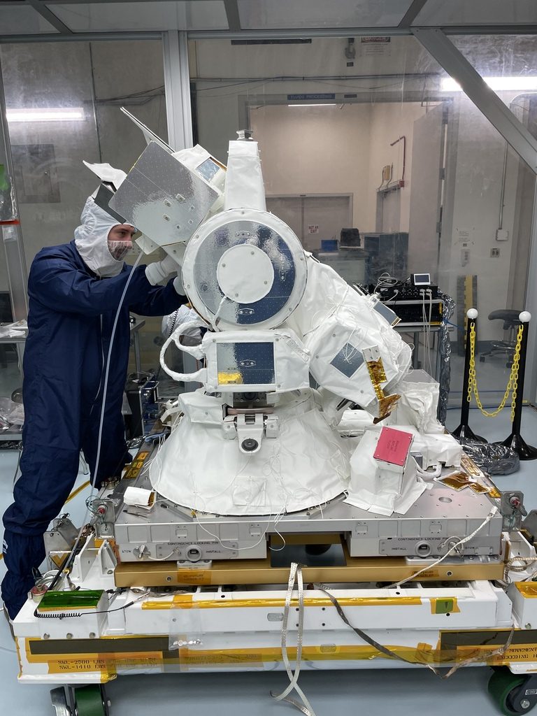 A person wearing a blue full body suit and a white head covering works on CODEX. CODEX is mostly white, and appears similar to a large, bulky telescope. It's on a platform.