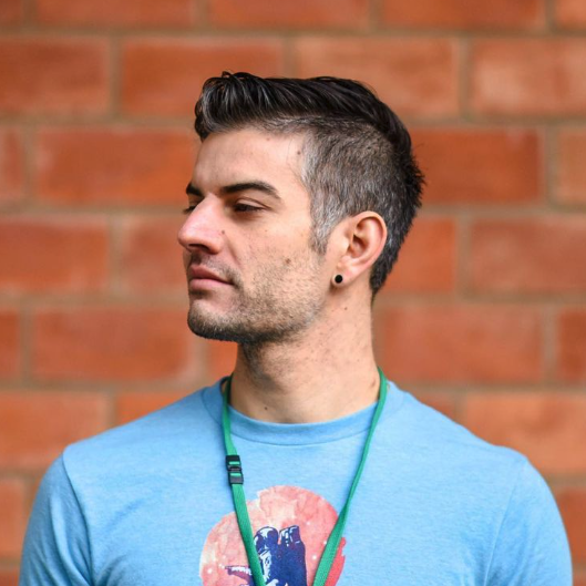 A white man in his late 30's, wearing a light blue shirt featuring an astronaut in front of a moon, stands in front of a brick wall and looks off to the left.