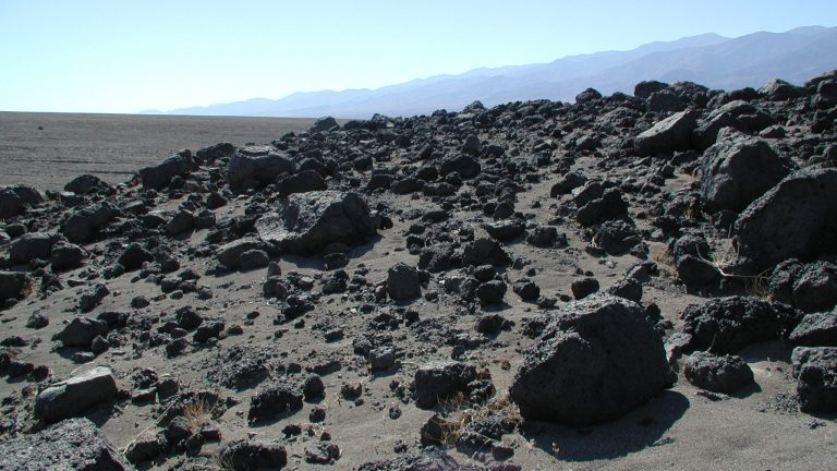 A dark grey, barren hillside covered in boulders of many sizes. In the background: distant blue mountain ridgelines under a clear sky.