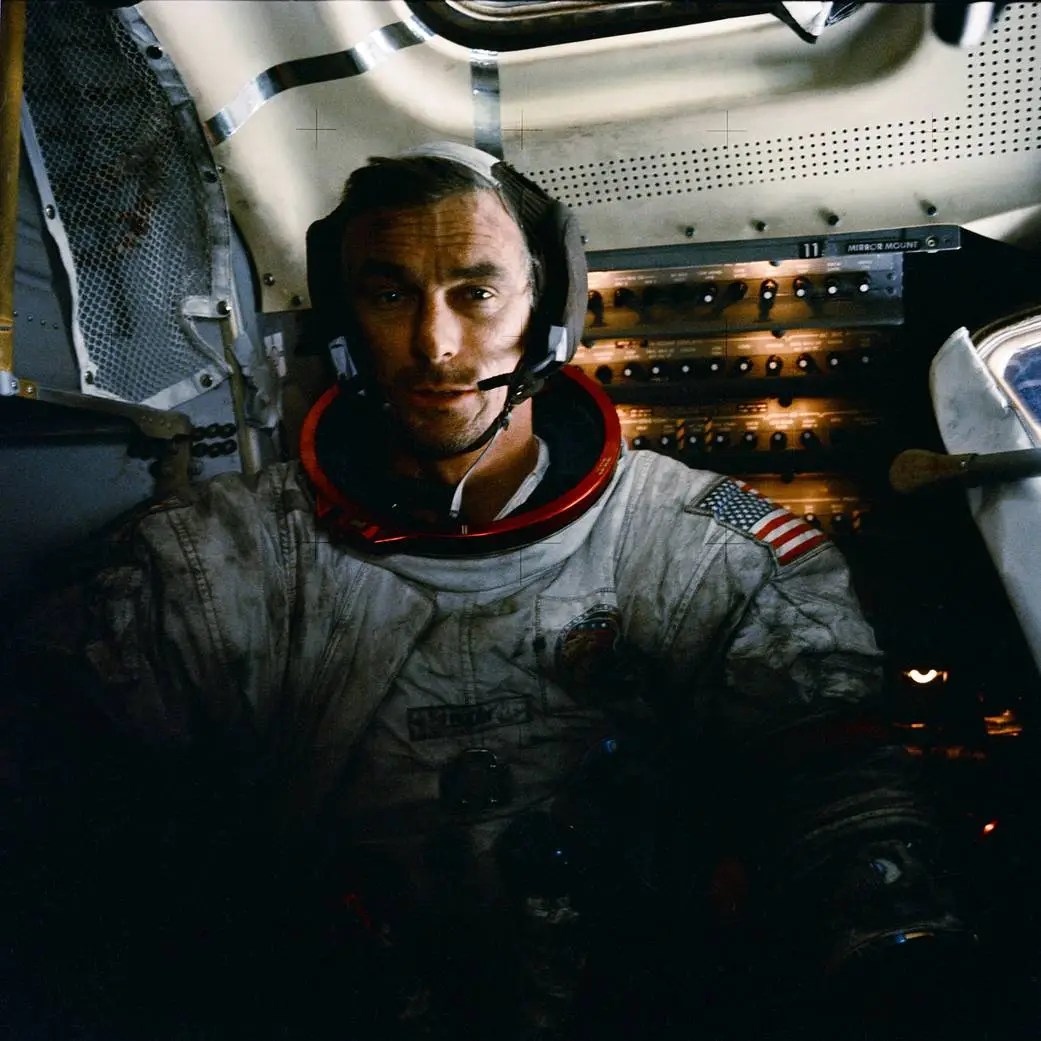 A person wears a bulky spacesuit that is shadowed on the bottom half. His suit and face are covered in dark smudges. He wears a headset with a microphone and his shoulder displays the American flag. Behind him, is a small space with controls and dim lights.