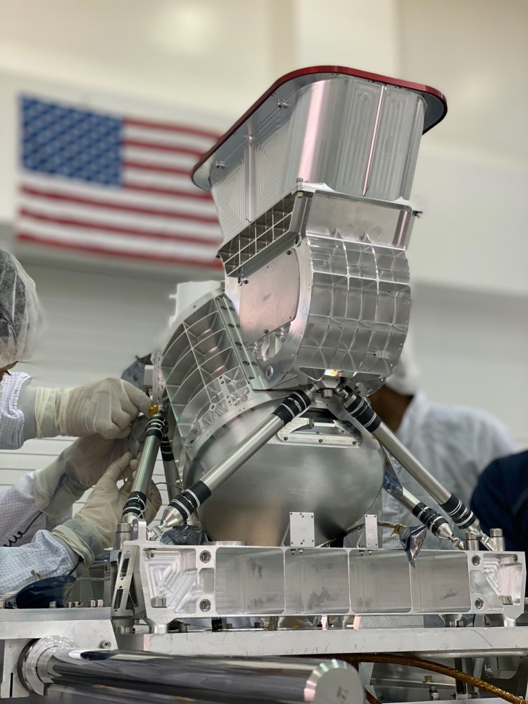 An engineer in protective clothing and gloves works on a silver-colored instrument with a red cap on top.