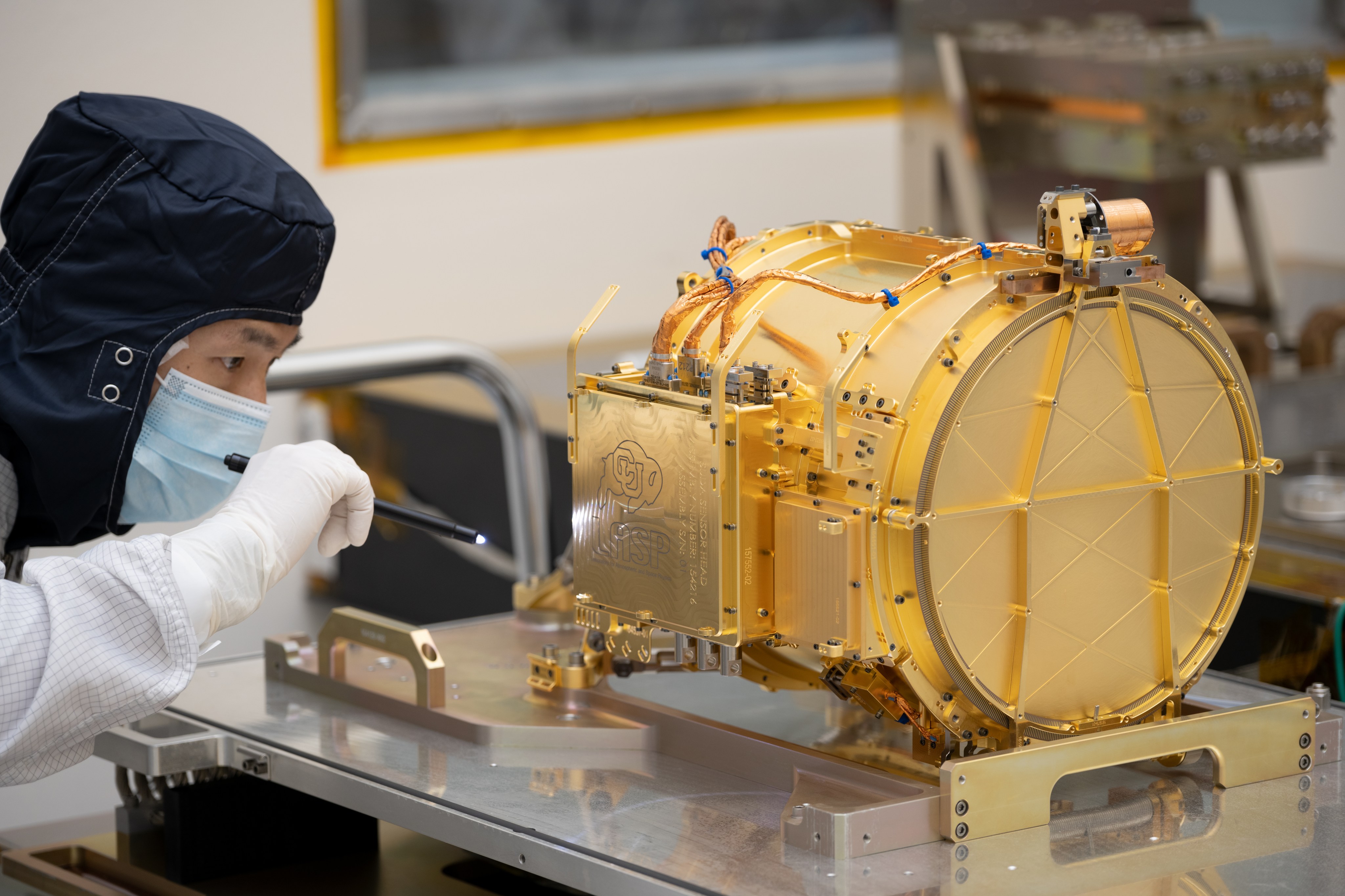 An engineer wearing a blue protective head covering examines the gold-colored, cannister-shaped SUDA instrument as it sits on a metal table.