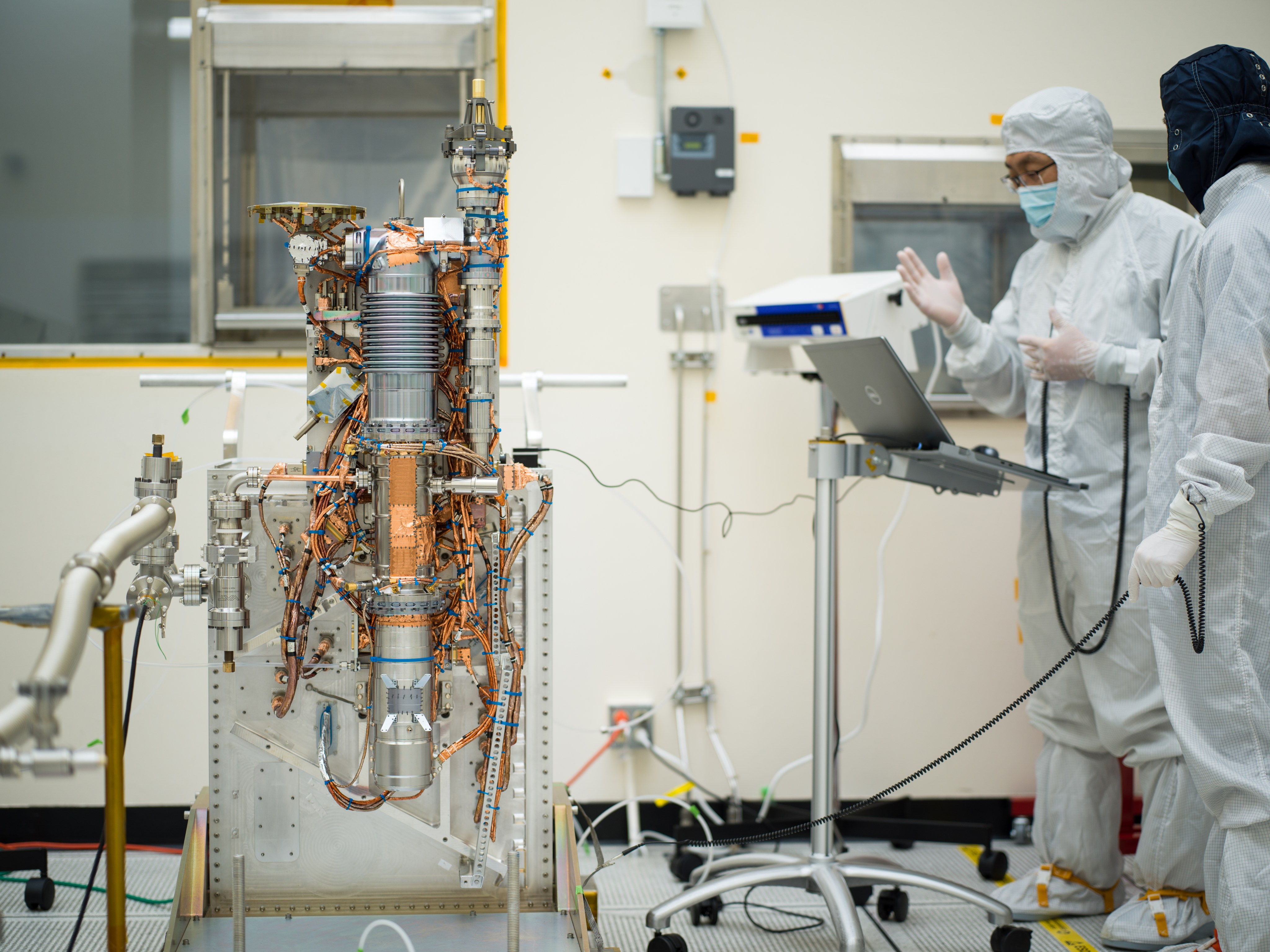 Two workers in protective clothing stand at a laptop on a stand as they inspect the MASPEX instrument sitting nearby. The instrument is on a stand making it about as tall as the workers. It has gold- or copper-colored wires secured by blue zip ties.