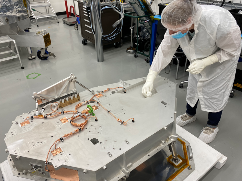 A worker in protective clothing and a blue face mask leans over a large, nearly hexagonal-shaped box with copper-colored wires running over it.