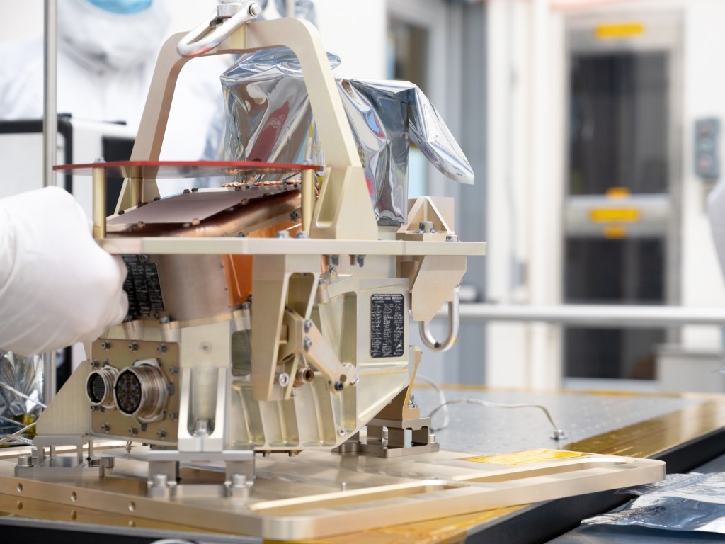 A cream-colored instrument sits on a workbench. A worker's gloved hand can be seen touching it.