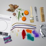 Several items are laid out on a gray table including a coloring page, a leaf, a sea shell, some green leaves, two oranges, a paint brush, a fork, a comb, three pipe cleaners, crayons, coins, toothpicks, a paperclip, features, and aluminum foil.