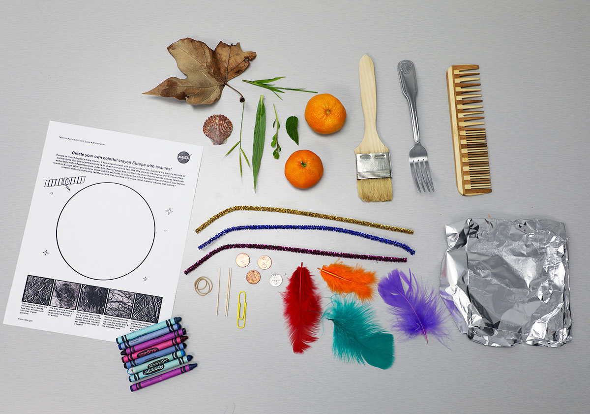 Several items are laid out on a gray table including a coloring page, a leaf, a sea shell, some green leaves, two oranges, a paint brush, a fork, a comb, three pipe cleaners, crayons, coins, toothpicks, a paperclip, features, and aluminum foil.