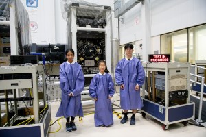 Two girls and a boy are wearing oversized smock in a test facility. A lighted sign says "Test in Progress."