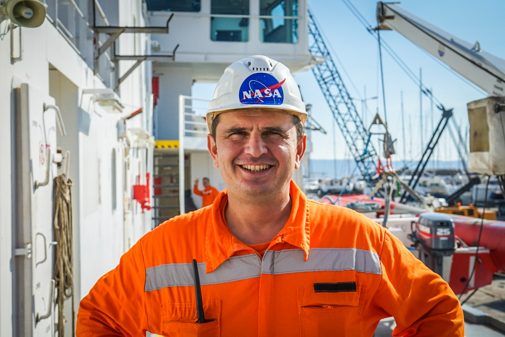 A smiling member of the crew wearing an orange jumpsuit and a white hard hat with a NASA sticker on it.