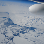 A white part of an airplane is shown in the sky over a patchy, icy part of Earth. A small bit of blue water can be seen between the glaciers and sea ice.