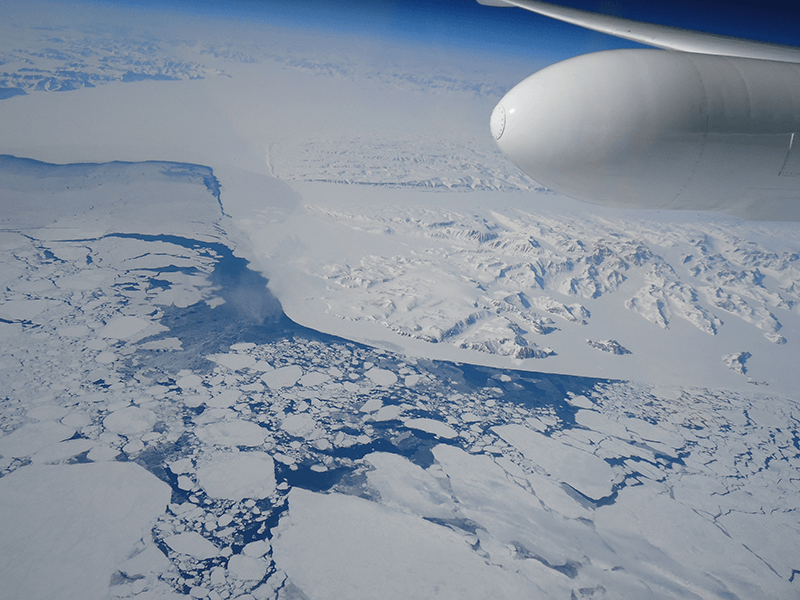 A white part of an airplane is shown in the sky over a patchy, icy part of Earth. A small bit of blue water can be seen between the glaciers and sea ice.