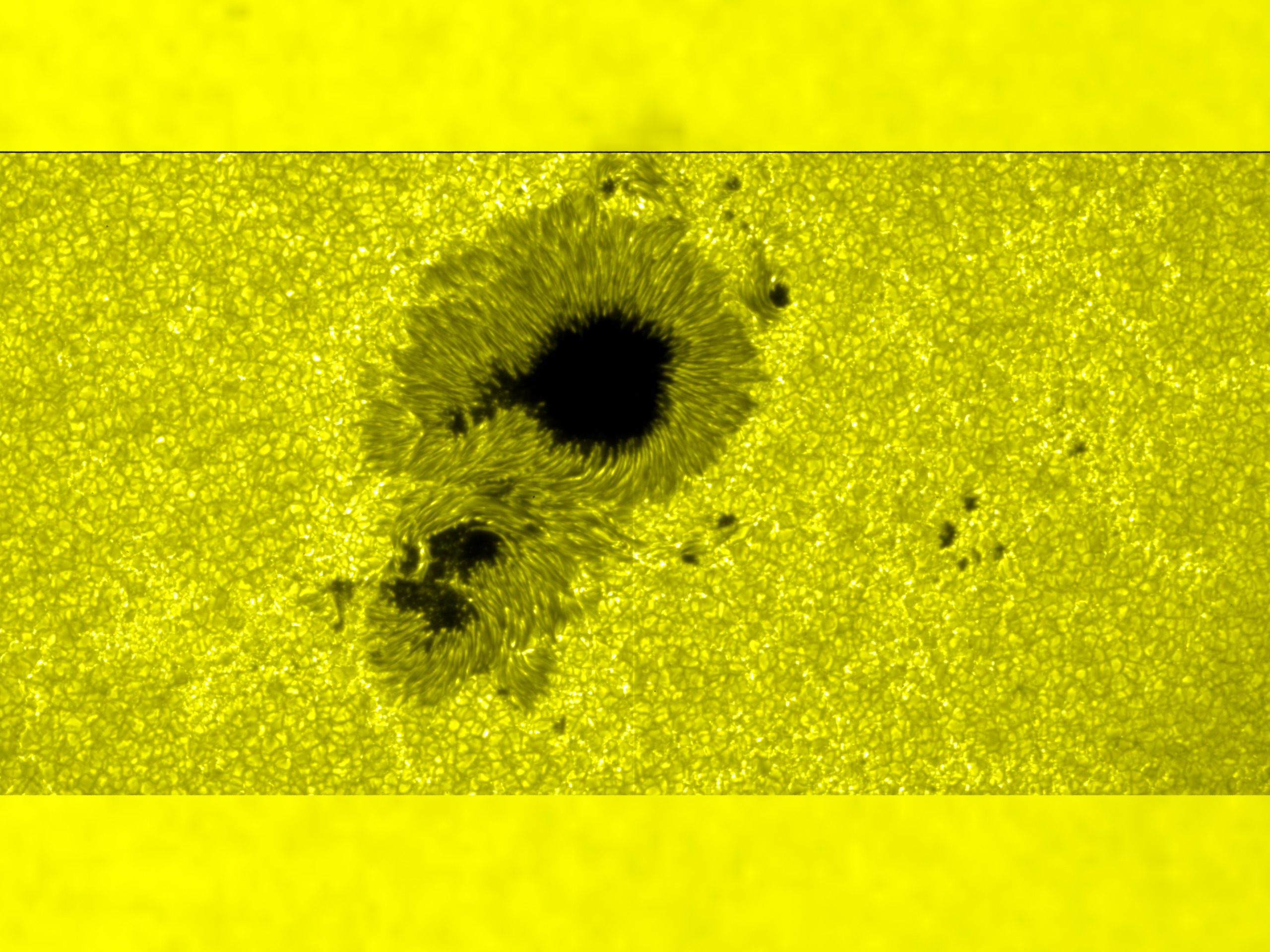 A sunspot seen in bright yellow. It appears as a few dark splotches, with lines of a darker yellowish gray that move outward from the splotches in every direction.