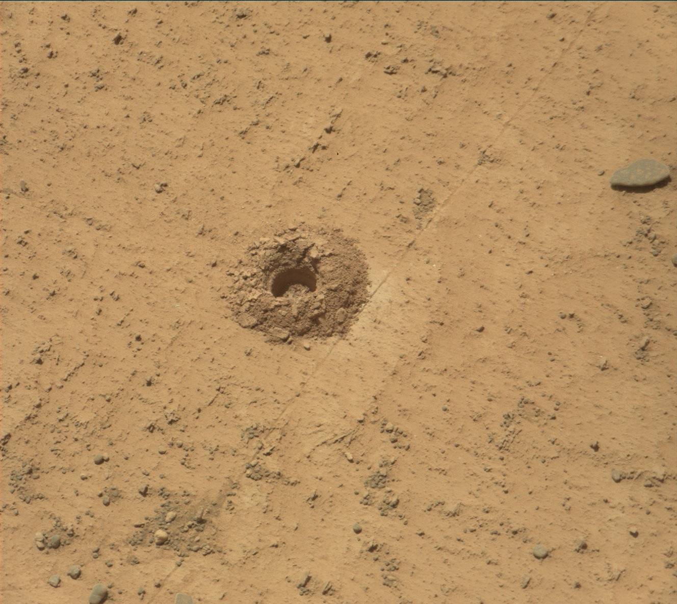 A photograph of a patch of gritty ground, mostly packed flat with scattered tiny sharp rocks and a few small ridges. In the center of the image, a shallow hole has been bored into the ground, and a pile of the excavated soil forms a mound around the hole.