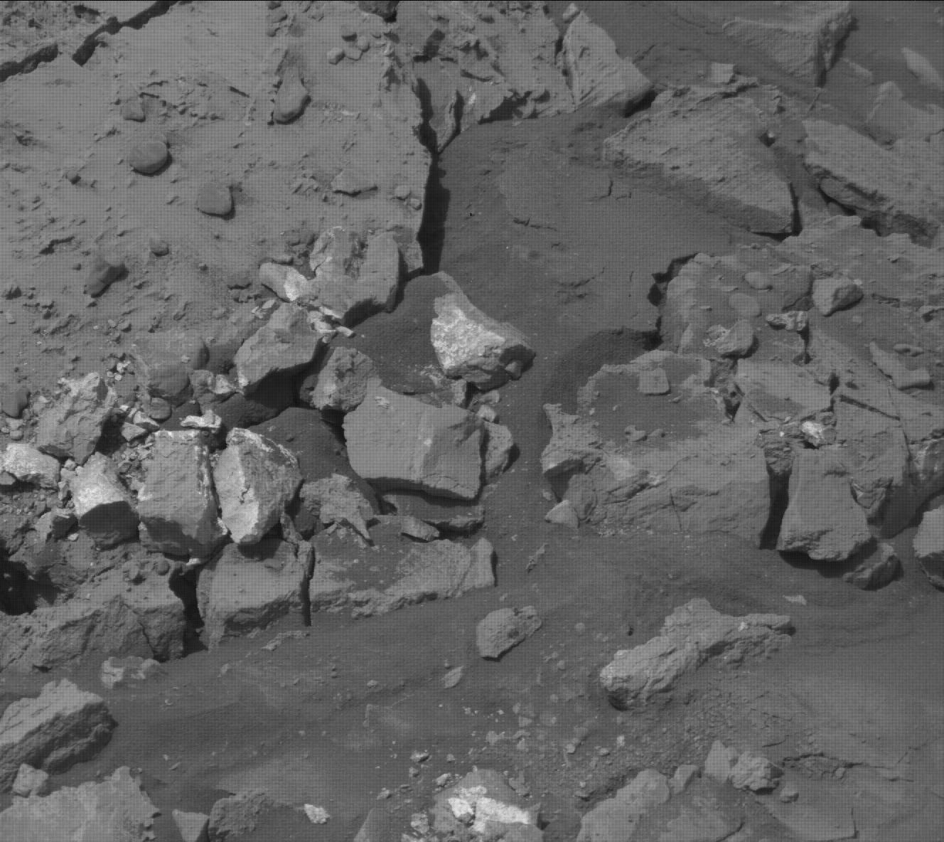 A grayscale photo of the Martian surface shows rocky terrain in shades of medium gray, with larger slabs and smaller rocks interspersed with areas of smooth soil, but several small-to-medium rocks stand out — in the middle-left and bottom parts of the frame — having surfaces much brighter and lighter-colored than anything around them.