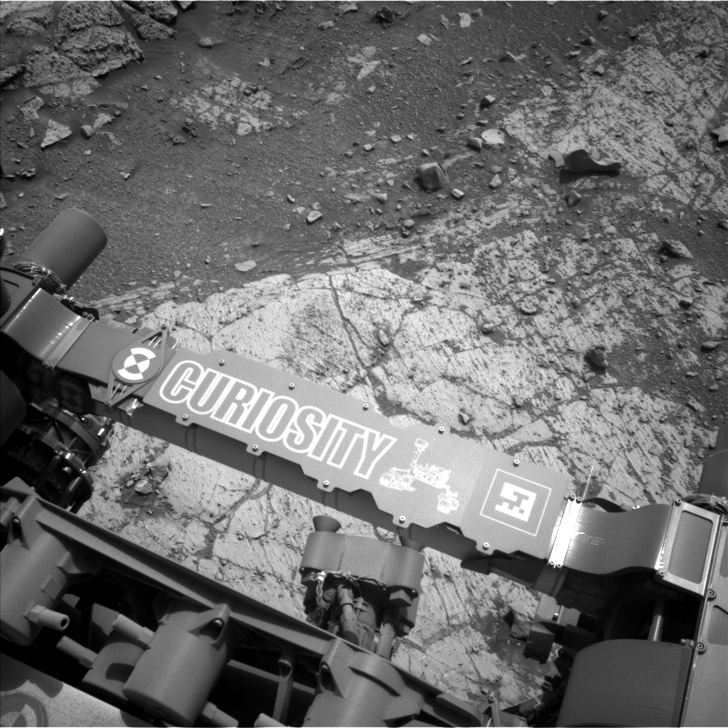 A grayscale photograph of the Martian surface from the Curiosity rover captures parts of the rover in the bottom third of the frame, including a crosspiece imprinted with its name and a line drawing of Curiosity. Ahead of the rover, on the right side of the frame, terrain consisting of large slabs of flat, light gray rock are criss-crossed with long, dark gouges, and sprinkled with small rocks. The left side of the frame is also flat, but with smaller slabs barely visible underneath the soil covering them. Beneath Curiosity, in a patch of ground visible between the rover’s body and the crosspiece, is a very light-colored rock, visible just below the “C” in Curiosity’s name.