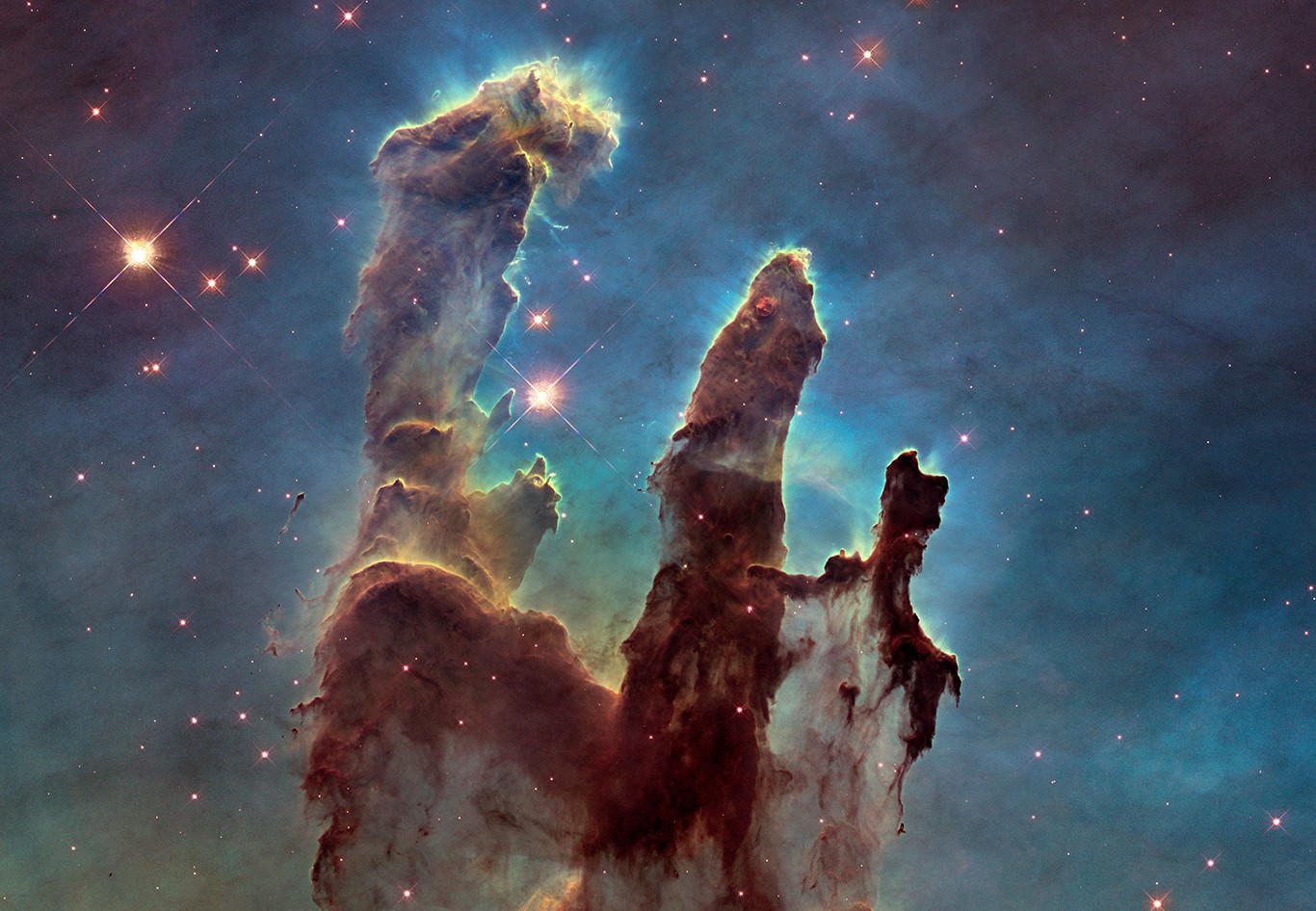 Three giant pillars of rusty colored dust and gas with a blueish green background dotted with stars.