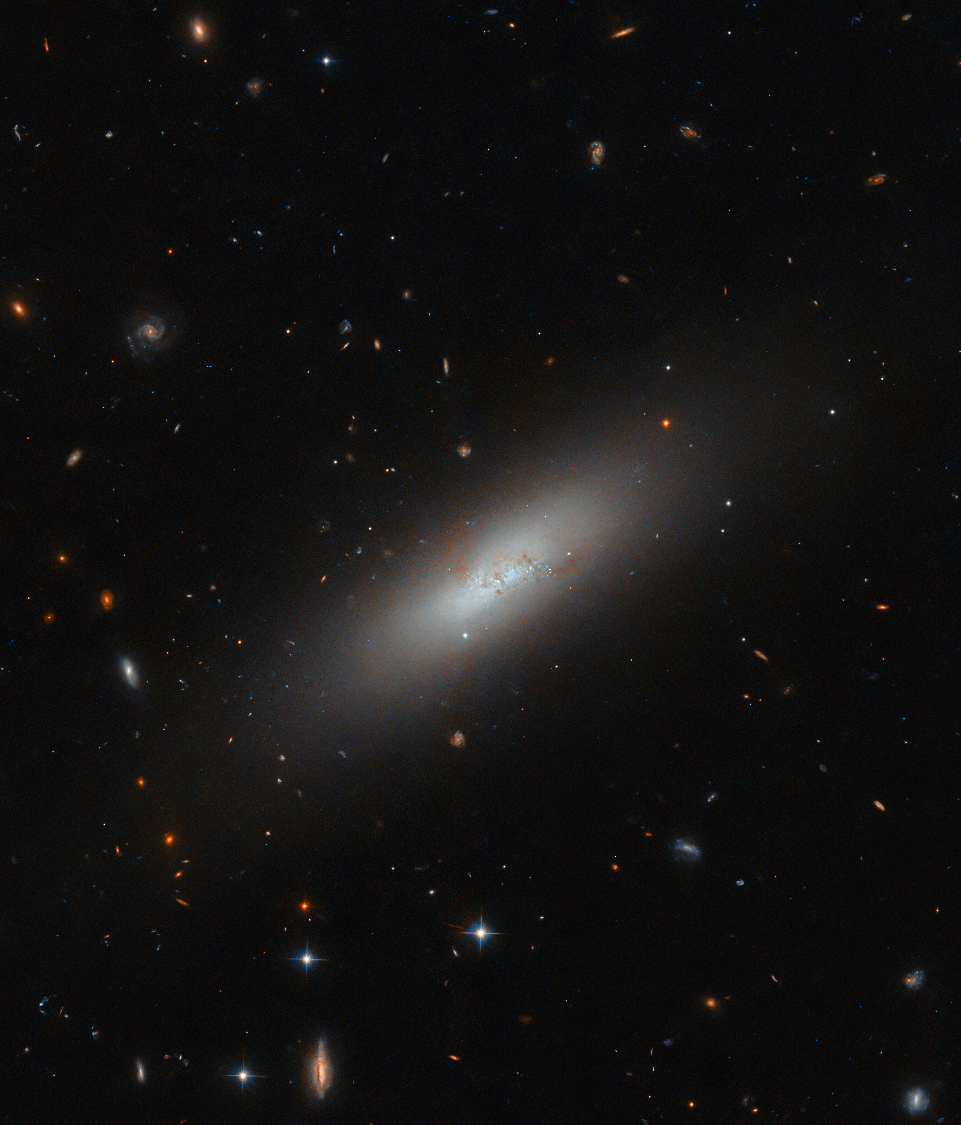 A relatively small, oval-shaped galaxy, tilted diagonally. Its center glows brightly and dims gradually to its edge. Its center also holds a few small, blue, glowing spots where stars are forming and dark wisps of dust that cross the galaxy. The image has a black background on which many background galaxies and foreground stars are visible.