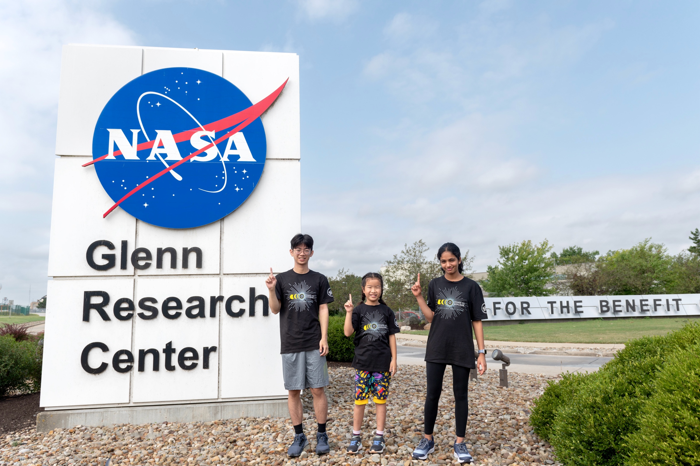 A boy and two girls are standing next to a NASA sign that says Glenn Research Center. They all have a finger up in a Number 1 sign.