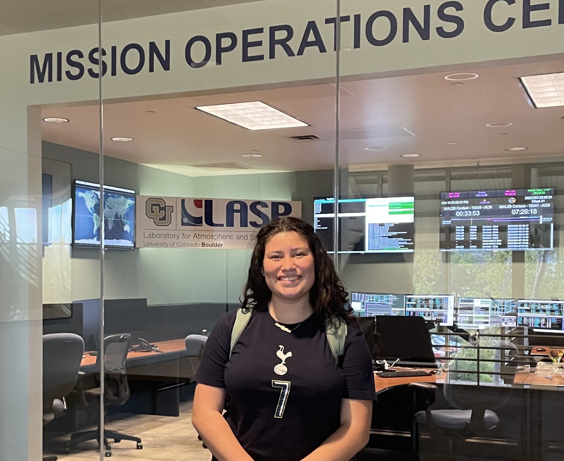 A young woman holds a water bottle in front of a doorway with the words "Mission Operations Center" overhead.