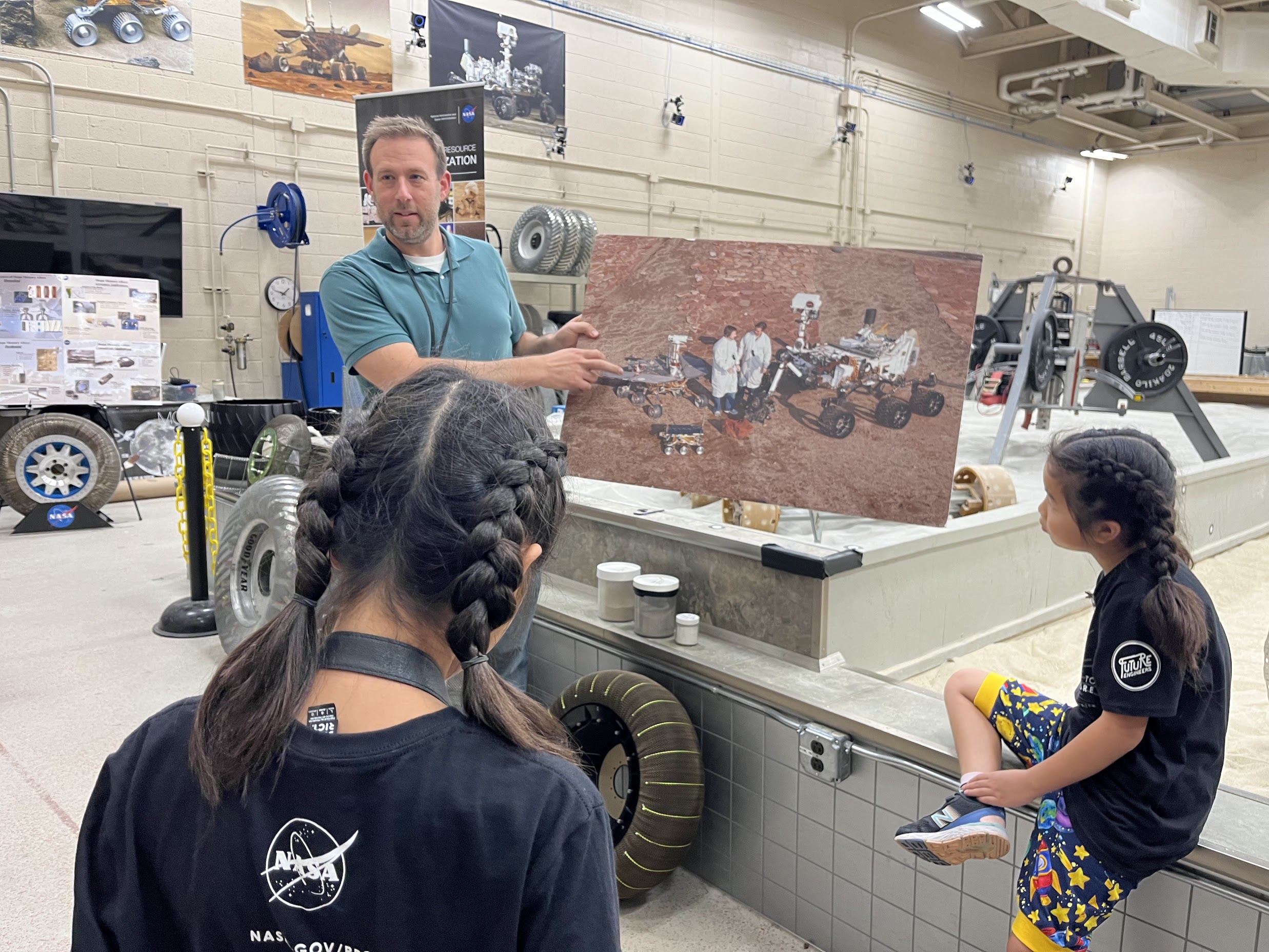 A man points to a picture of three Mars rover ranging in size from a child's wagon to an SUV. Two girls are listening to him talk.