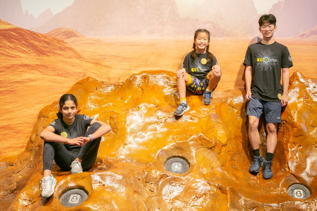 Two girls and a boy sit on a simulated display of the Martian surface.