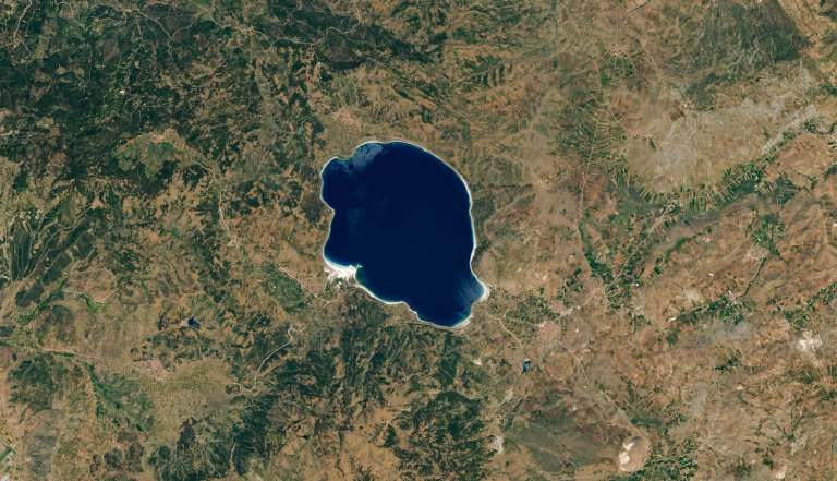 Satellite view of a deep blue lake surrounded by a green and brown landscape. The lake’s edge is clearly visible as a narrow band of light-colored material.