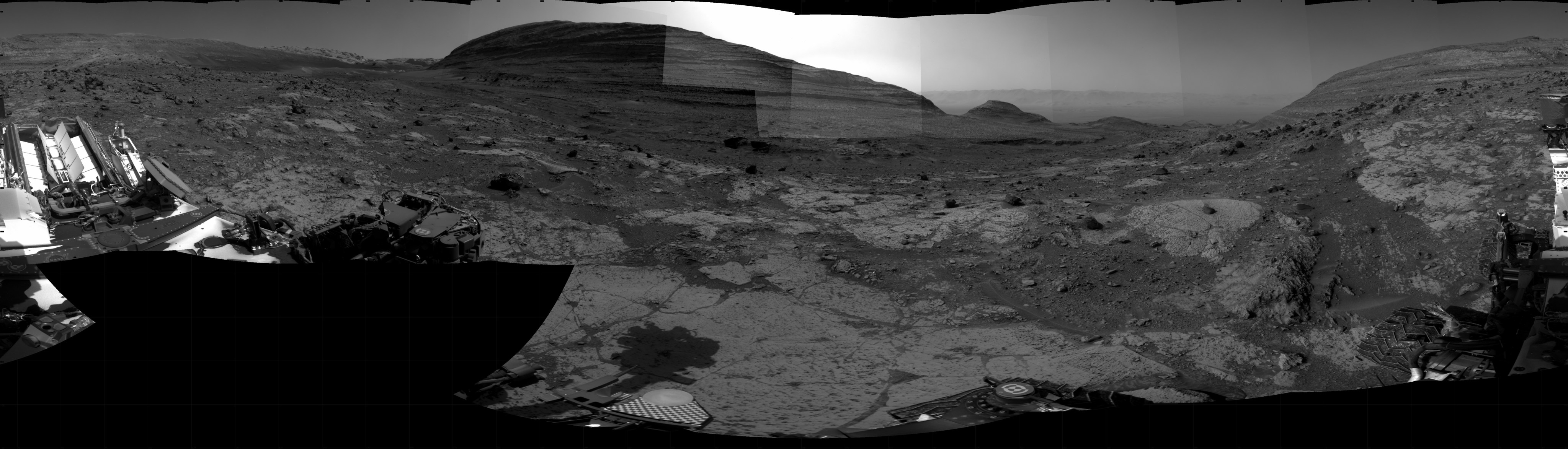 A grayscale photo panorama of the Martian surface shows an area of flat ground in the foreground, with some smooth and some rocky sections interspersed, and two tall buttes in the background, one at the right side of the image and one dominating the center. The center one is brightly backlit by glowing sunlight. Portions of the Curiosity rover are visible at the far left, bottom, and far right of the frame.