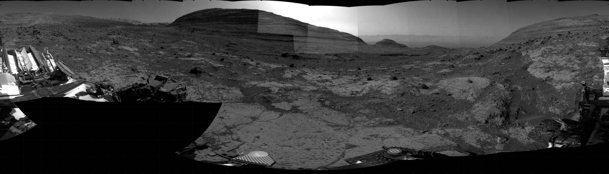 A grayscale photo panorama of the Martian surface shows an area of flat ground in the foreground, with some smooth and some rocky sections interspersed, and two tall buttes in the background, one at the right side of the image and one dominating the center. The center one is brightly backlit by glowing sunlight. Portions of the Curiosity rover are visible at the far left, bottom, and far right of the frame.